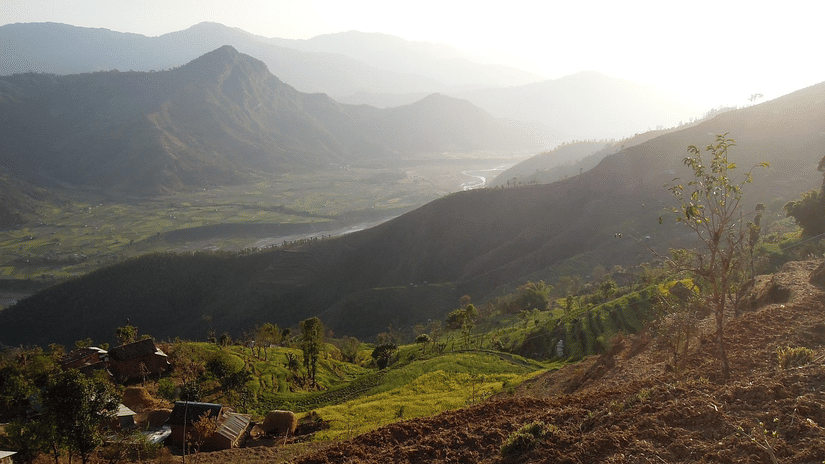 A wide landscape view of rolling hills and mountains under a bright, hazy sky with sunlight hitting the foreground.