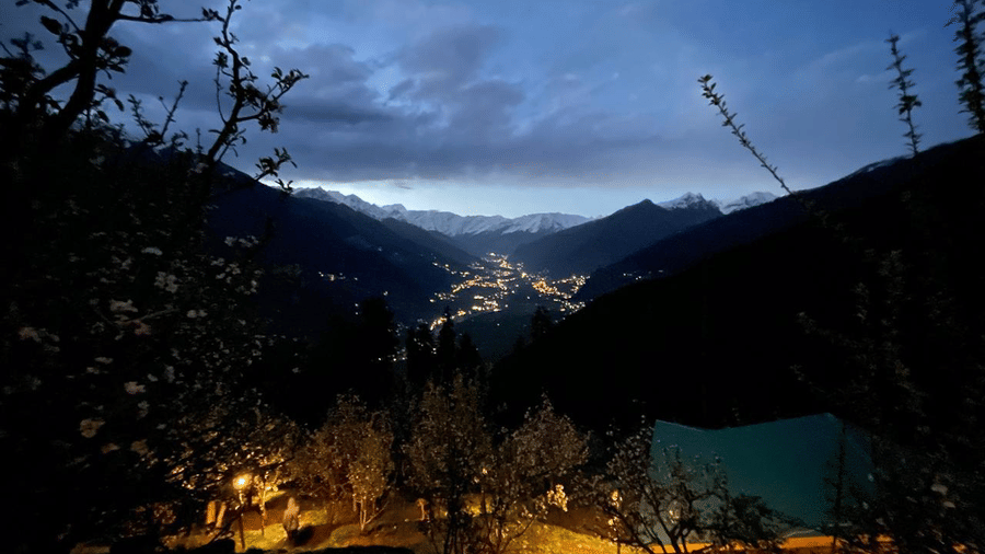 A late evening view from Amara Upepo - The Sky Village, Manali, looking down into the mountain valley where the town lights glow brightly, contrasting with the deep blue.