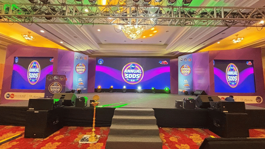 A large hall with rows of sofas facing a lit stage, patterned carpeting, and bright ceiling lighting.