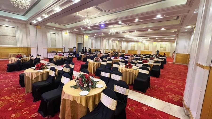  A banquet hall at Amer Greens is set for an event with cocktail tables draped in gold linen, surrounded by chairs in black covers and gold sashes, on a red and gold patterned carpet.