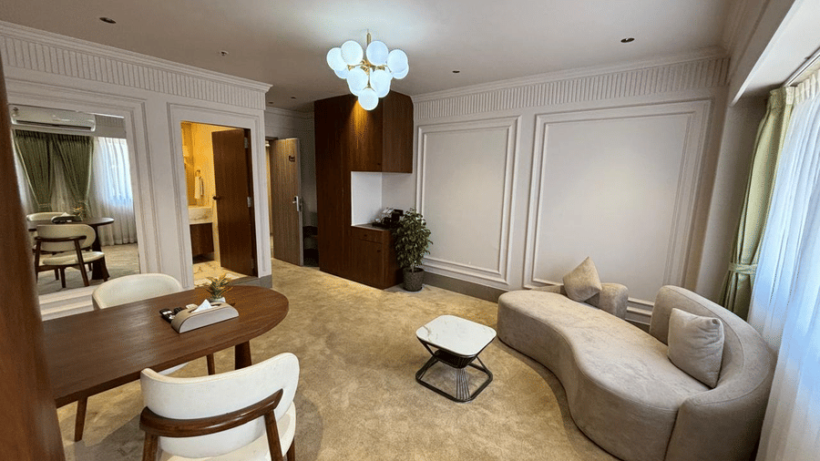 Modern living area of a luxury hotel suite at Biznotel by Pride Surat featuring a curved sofa, dining table, and contemporary chandelier.