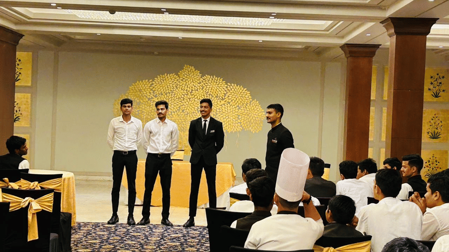 A line of individuals stands facing an audience during a gathering inside Clarks Group of Hotels. People sit at tables.