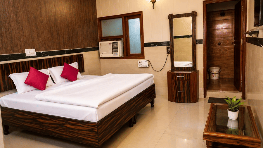 Bedroom interior of Hotel Goverdhan, Agra, featuring king size bed with white linen, 2 pillows, wooden headboard, window air conditioner, dressing table, mirror, table, and plant.