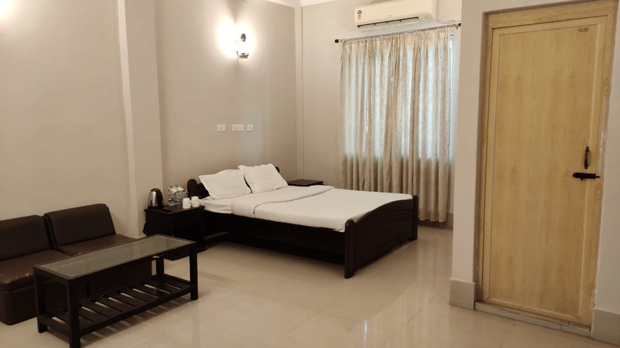 A room at Lake Side Resort Neermahal, featuring a bed, bedspreads, a pillow, a sofa, a table, and a window covered with curtains.