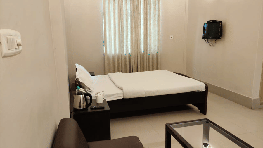 A room at Lake Side Resort Neermahal, featuring a single bed, bedspreads, a pillows, a sofa, a table, a window covered with curtains, and wall-mounted lamps.