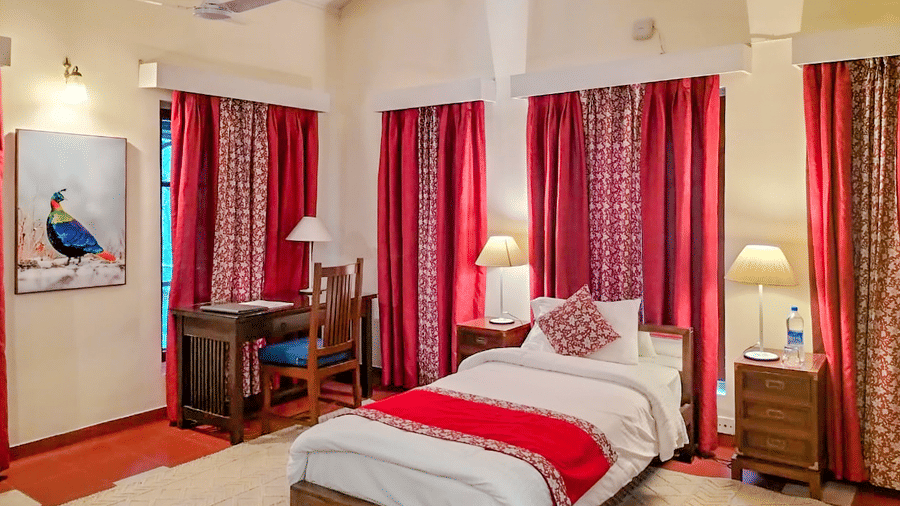 Spacious heritage bedroom with a wooden double bed soft lighting and rich red curtains creating a warm and restful ambience at Neemrana’s Glasshouse on The Ganges with traditional furnishings high ceilings and a calm indoor atmosphere ideal for relaxation