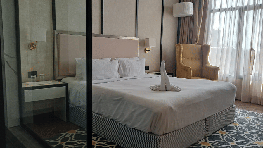 Guest room at Polo Hotel, Agartala showing the entry door, full-length mirror, glass-partitioned bathroom and king bed with patterned rug.