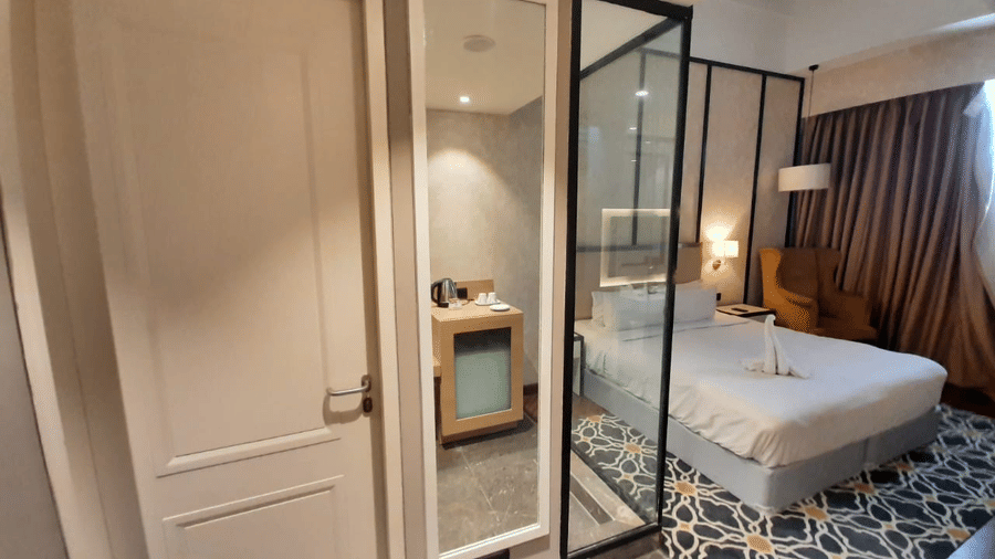 Guest room interior at Polo Hotel, Agartala with white entry door, full-length mirror, glass partition, double bed and yellow armchair.