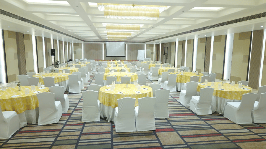 Spacious banquet hall at The Piccadily featuring yellow-clothed round tables, white chairs, a projection screen, and bright overhead lighting for corporate events.