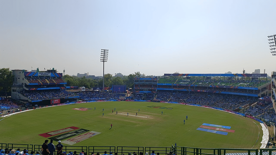 Panoramic cricket stadium with lush green outfield and crowd during day match.