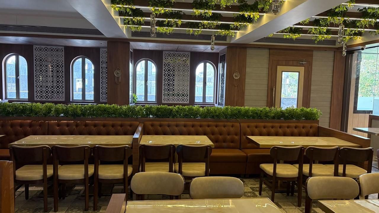 A brightly lit restaurant area with wooden tables and chairs, featuring arched windows and some greenery - Melior Inn