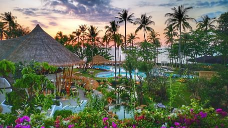 Tropical resort at sunset with lush vegetation and thatched-roof structures.