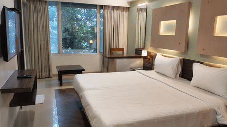 View of a room at Hotel Mumbai House with a king bed, wooden nightstand, large window, and lush greenery beyond.