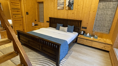 Megha Resort & Villas, Dehradun Standard Room, featuring a dark timber bed frame, timber-clad walls, a smooth light floor, and an internal wooden staircase leading to another level.