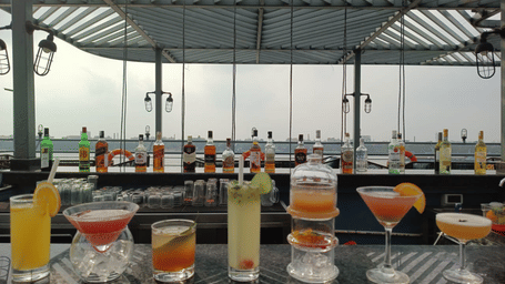A bar counter with various alcoholic bottles on top and cocktail glasses in front, with a river and a bridge in the background under a roof structure.