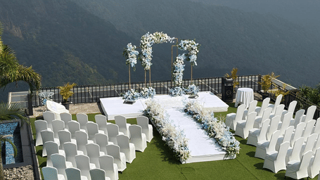 Outdoor wedding ceremony setup at Polo Resort Cherrapunji with white chairs, floral arch and aisle on grass overlooking misty green mountains.