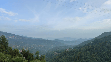 A scenic view of lush green mountains under a blue sky with scattered clouds at Sukama Resort & Spa, Mukteshwar.