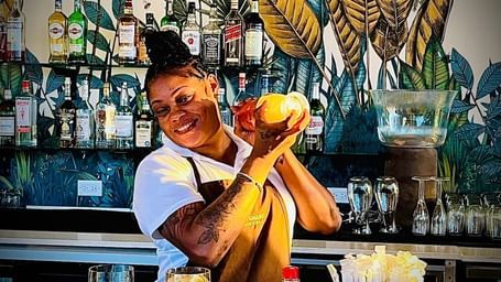 A bartender is shaking a cocktail shaker behind a bar with bottles and tropical-themed decor in the background.