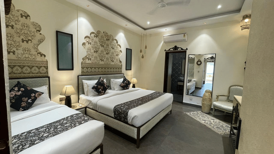 2 King size beds in a luxury suite at Beelwa Palace, Jaipur, featuring wall decor, bedside lamps, a mirror, and an armchair.