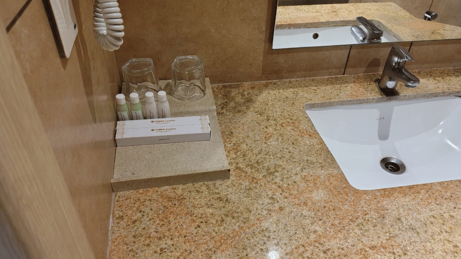 Washroom of the Standard Room at Cholaa Dynasty, Kumbakonam featuring a sink with faucet and some ointment tubes wit two glasses kept aside.