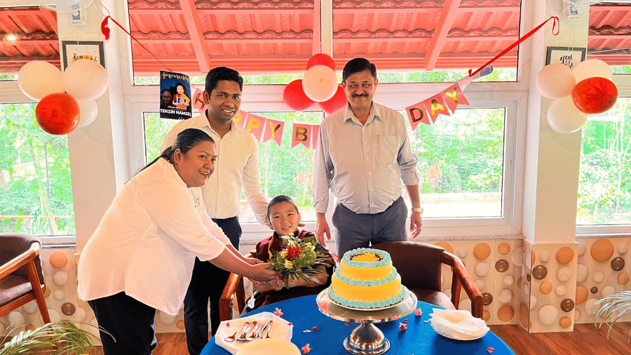 A family celebrating a birthday indoors, standing around a blue table with a yellow cake and balloons, with a red-tiled roof visible at Coorg Orange Blossom Resort and Spa.