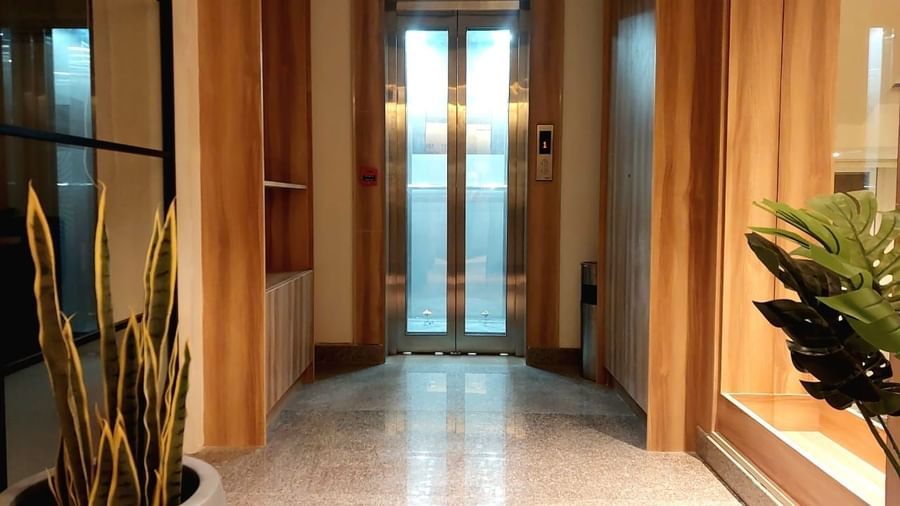 An elevator with its door closed at the end of the corridor at Hotel Raai, Hosur. The corridor has a few flower pots like snake plant and monstera plant.