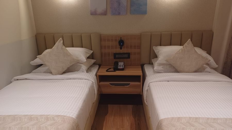 Premium Room at Hotel Paai, Hosur featuring twin beds placed at two corners of the room. Above each bed there are lights on the wall. Between the beds there is a bedside table with a telephone and socket. 