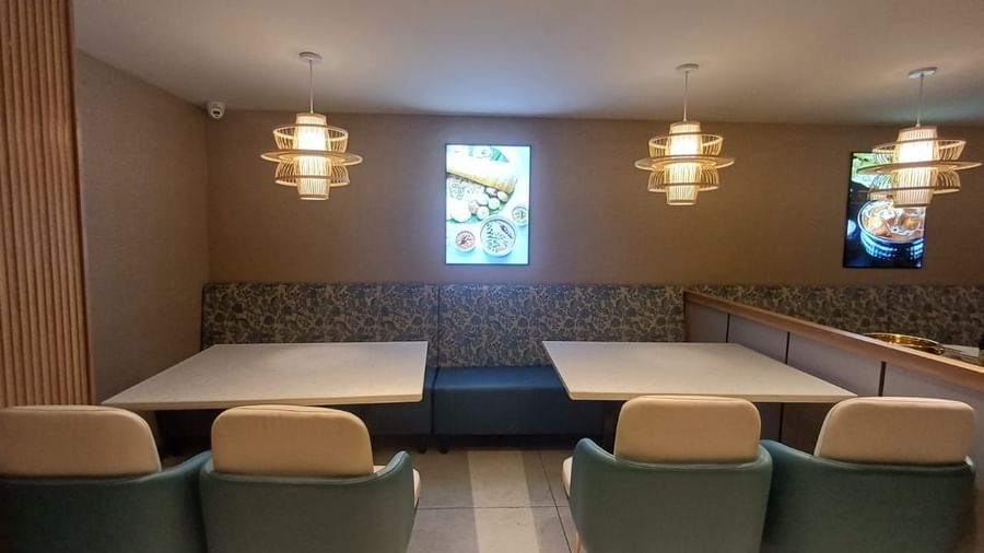 A restaurant at Hotel Raai, Hosur featuring sets of tables and chairs, lights hanging from the ceiling and a digital frames hanged on the wall. 