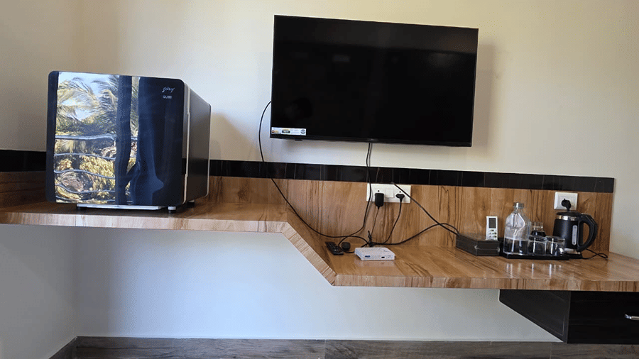 corner of the room showing a wall mounted TV, a mini fridge, and a wooden shelf with tea and coffee supplies.