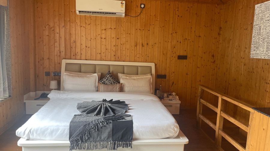 A wood-panelled bedroom with a white bed contrasts with a dark grey blanket and pillows at Megha Resort & Villas, Dehradun.