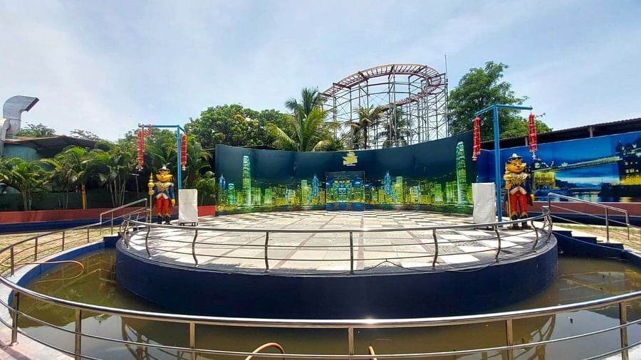 A circular stage surrounded by water, featuring decorative sculptures and a backdrop of amusement park rides and trees.