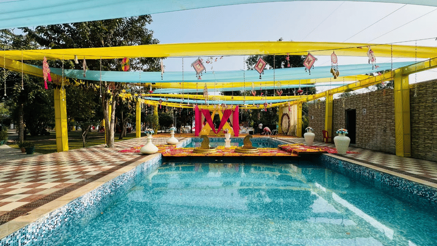 A large, covered outdoor area with a swimming pool in the foreground, yellow and blue draped fabric overhead, and a decorated ceremony structure visible in the distance | Nandan Resort