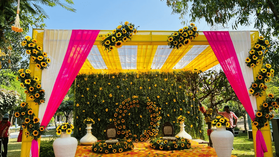 A brightly lit wedding stage in Bhopal, decorated with a canopy of yellow and pink fabric, featuring a dense floral backdrop and decorations on the ground | Nandan Resort