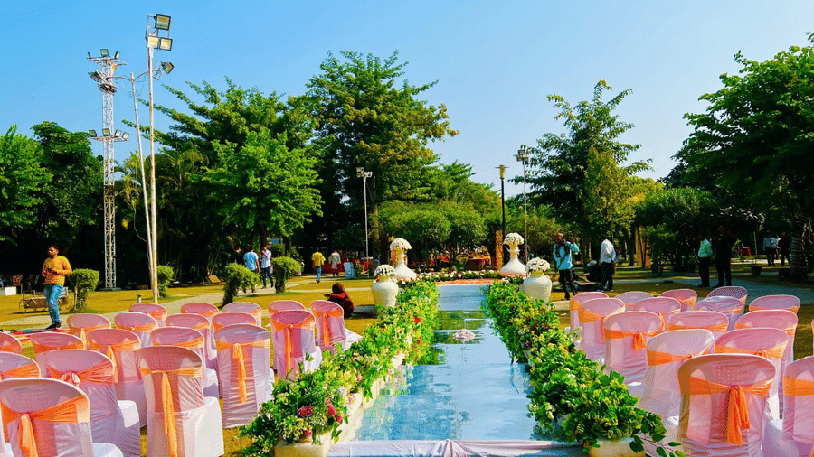 An outdoor wedding ceremony setup featuring an aisle lined with white chairs and peach-coloured sashes leading towards a floral arch, with a narrow water feature running down the centre | Nandan Resort