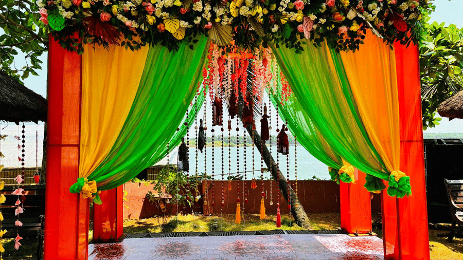 Wedding mandap decorated with orange and green drapes, flowers, and hanging garlands at Paradise Lagoon Resort, Udupi.