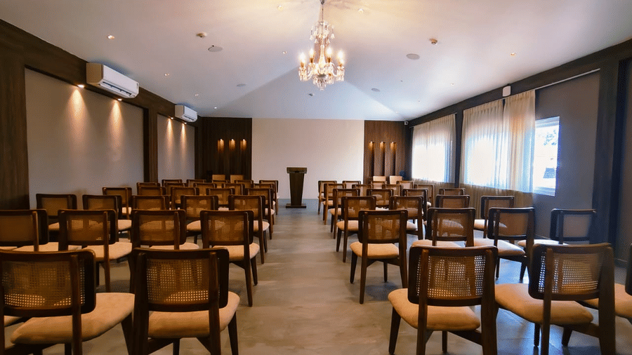 A conference room featuring rows of wooden cane-back chairs facing a wooden podium under chandeliers at Savyasaachii Hotel.