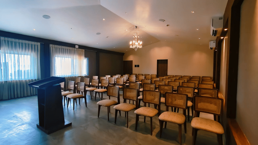 Rows of wooden chairs facing a podium in a brightly lit hall with chandeliers at Savyasaachii Hotel.