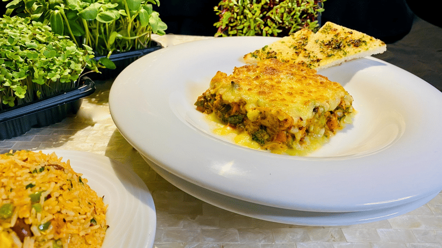 White plate with a slice of Italian vegetable lasagna topped with melted cheese and a garlic bread piece, with micro greens in the background.