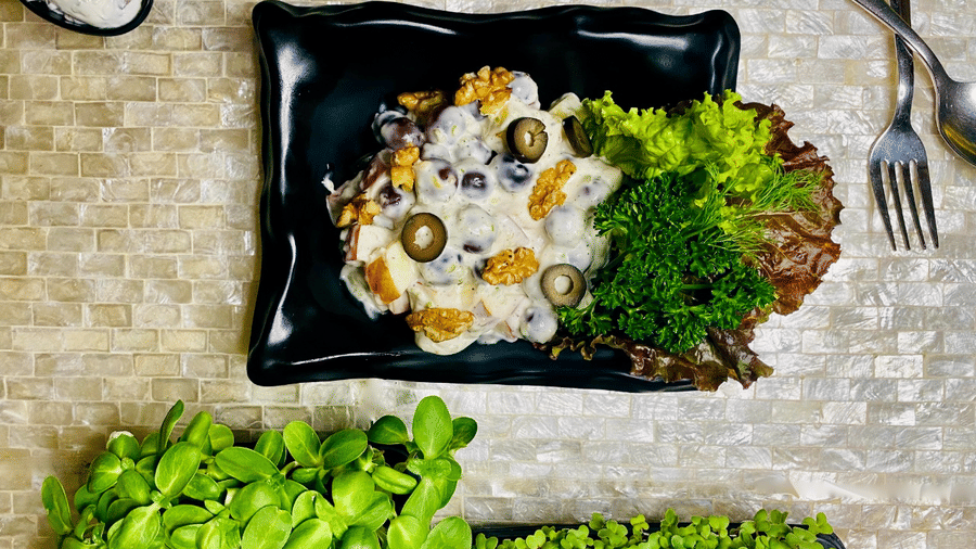 Black plate of Waldorf salad with grapes, apple, walnuts, olives and creamy dressing on lettuce, viewed top-down beside cutlery and micro greens.