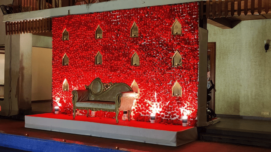 A decorated wedding stage at Vedic Village Spa Resort featuring a bold red floral backdrop hanging lanterns a sofa seating arrangement and warm ambient lighting in an evening setting.