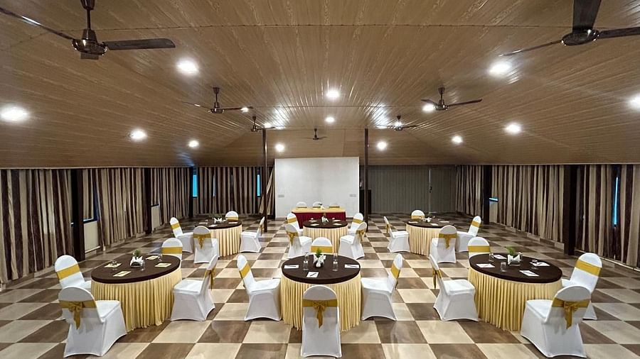 An indoor event space is arranged with round tables set with ceiling fans and lighting visible | Ballroom | Woodstock Resorts, Coorg
