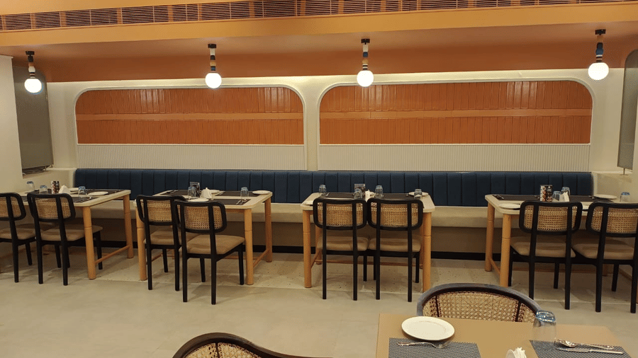 Dining tables with cushioned seats attached to wall on one side and chairs on the other side of the tables with overhead warm lighting at hotel Manorath in Lords Eco Inn, Belagavi.