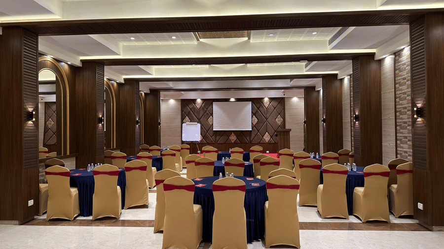 A view of a spacious contemporary hall featuring multiple decorated round tables and chairs around it facing a mounted screen at the backdrop at Shyanu Lords Eco Inn Airoli.