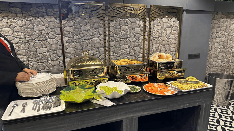 A buffet spread on a table set against a brick wall showcasing different food and fruits with a partially visible staff on the left at Shyanu Lords Eco Inn Airoli.