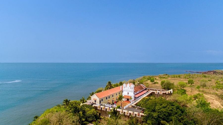 A panoramic view of the coastline at Fort Tiracol, Goa, featuring a white building with a red roof situated on a green headland overlooking the deep blue ocean under a clear sky.