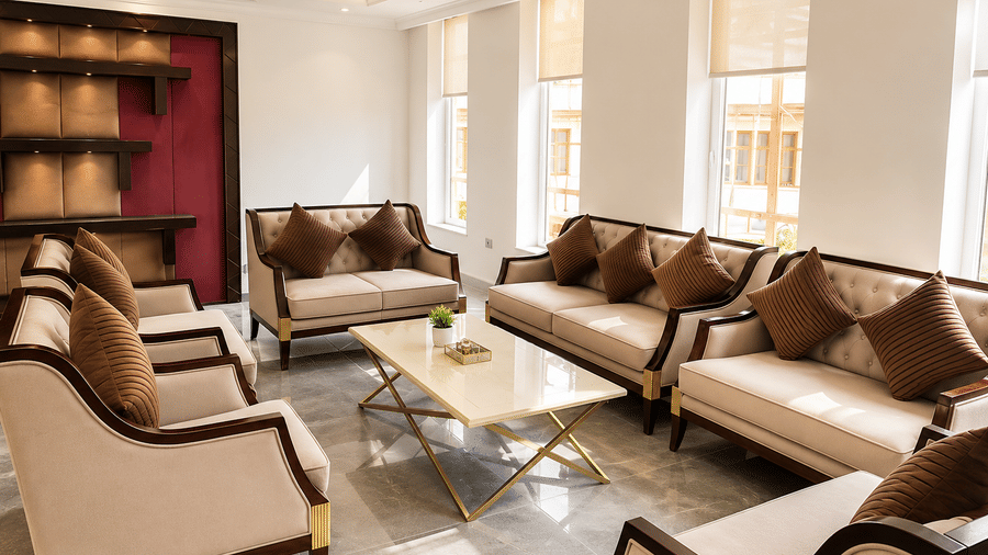 Hotel lounge with beige sofas and armchairs set around a glass coffee table, tall windows and a dark red bookshelf in the background nearby.