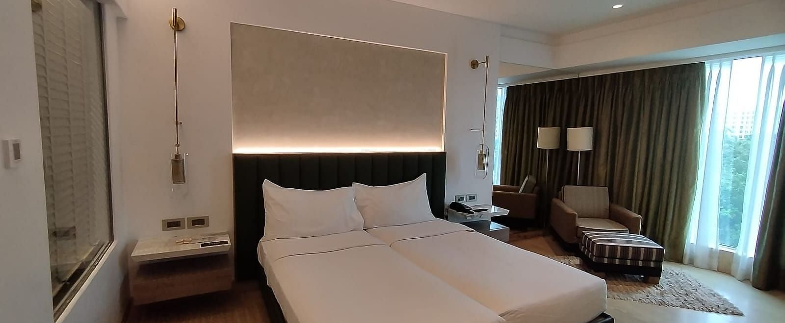 A warmly lit executive bedroom in Mirage Hotel, featuring twin beds, decorative head-rest, lounge chairs, and a large wall to wall window with partially open drapes.