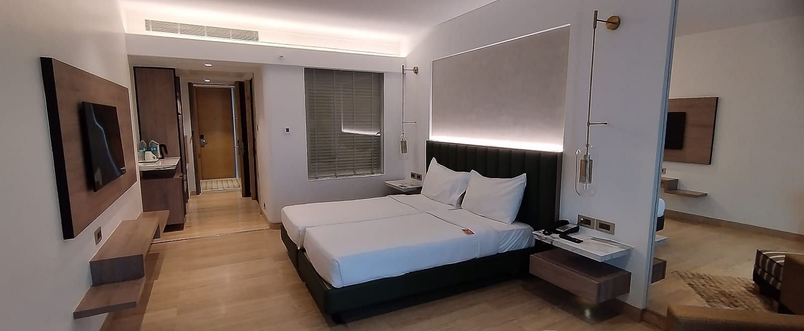 A wooden floored executive bedroom in Mirage Hotel, featuring twin beds, a large mirror, lounge chairs, and an entertainment unit.