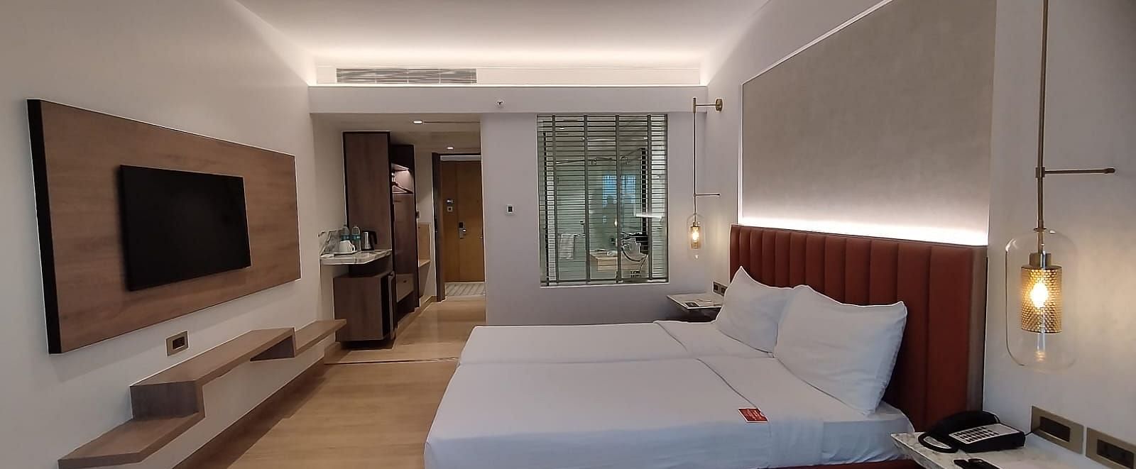 A well-lit, wooden floored, executive room in Mirage Hotel, featuring twin beds with decorative head-rest and a wooden panelled entertainment unit.