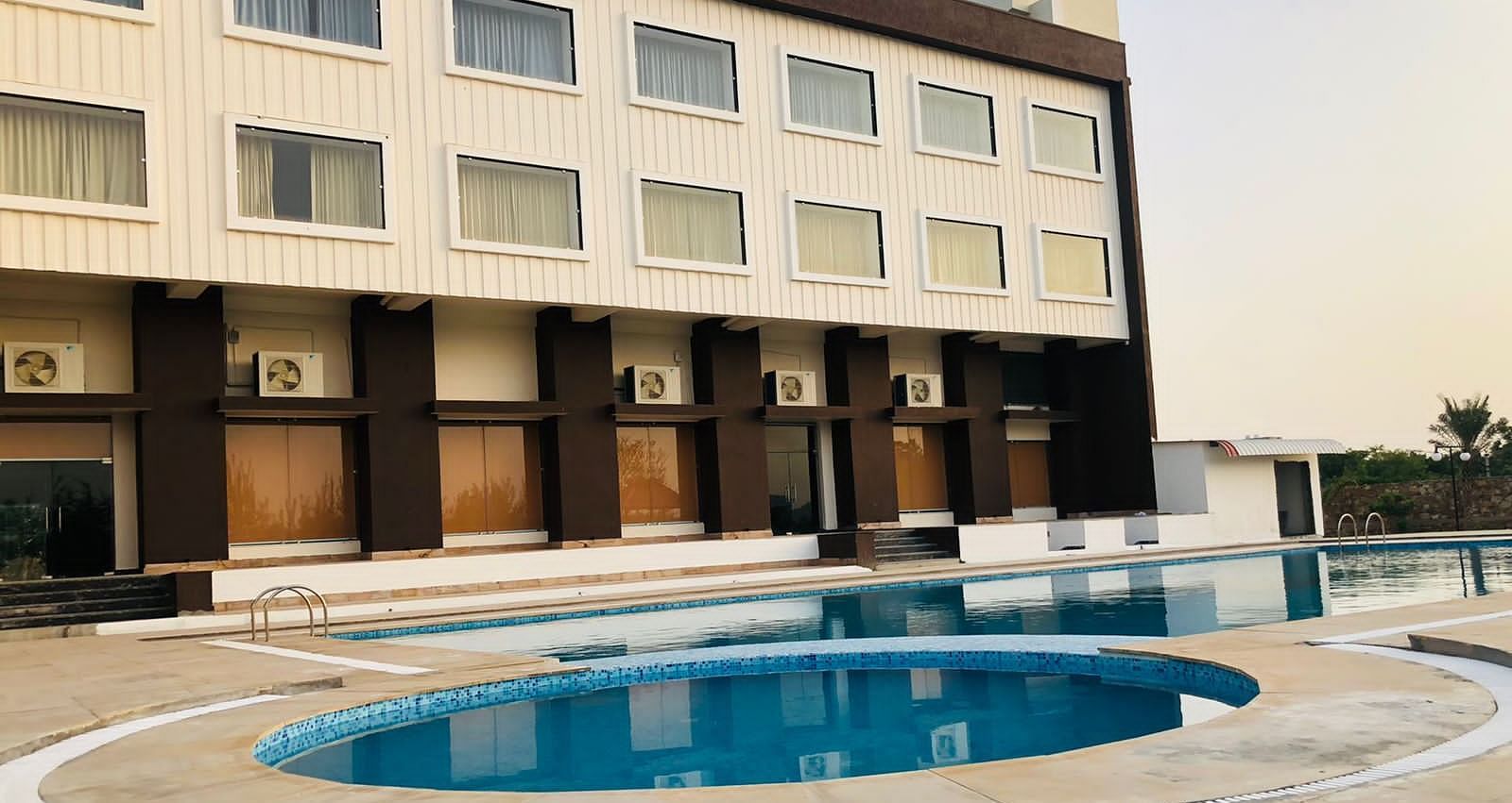 The exterior of the Bamboo Saa Mulberry Resort, Pushkar, featuring a multi-story building with a light-coloured facade with a swimming pool is visible in the foreground.
